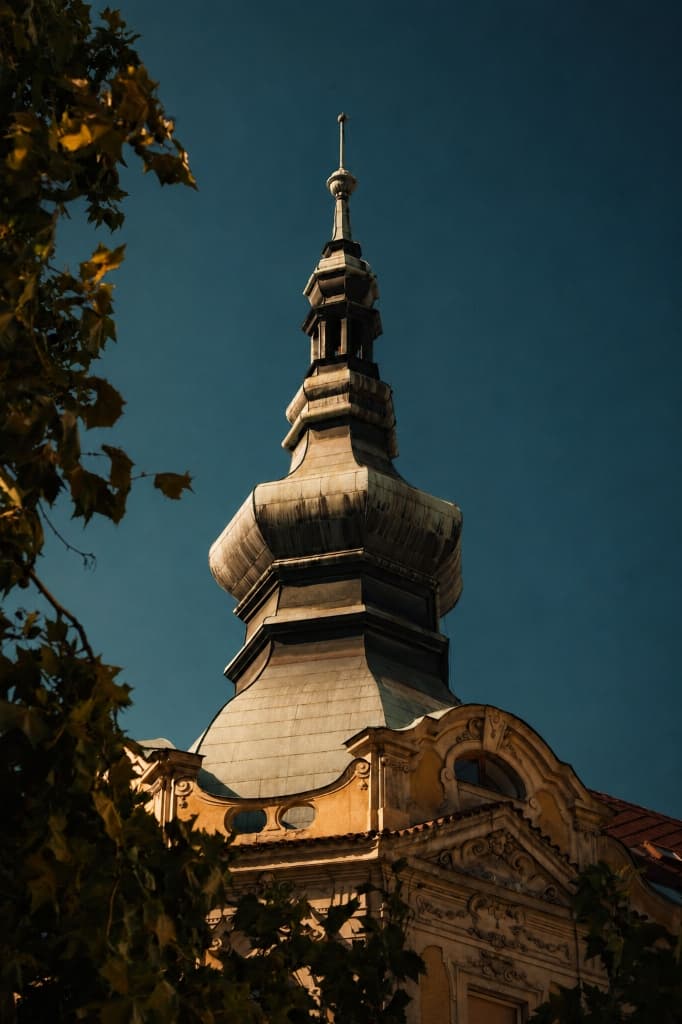 Historic Prague baroque spire and ornate architectural roofline framed by foliage under deep blue sky